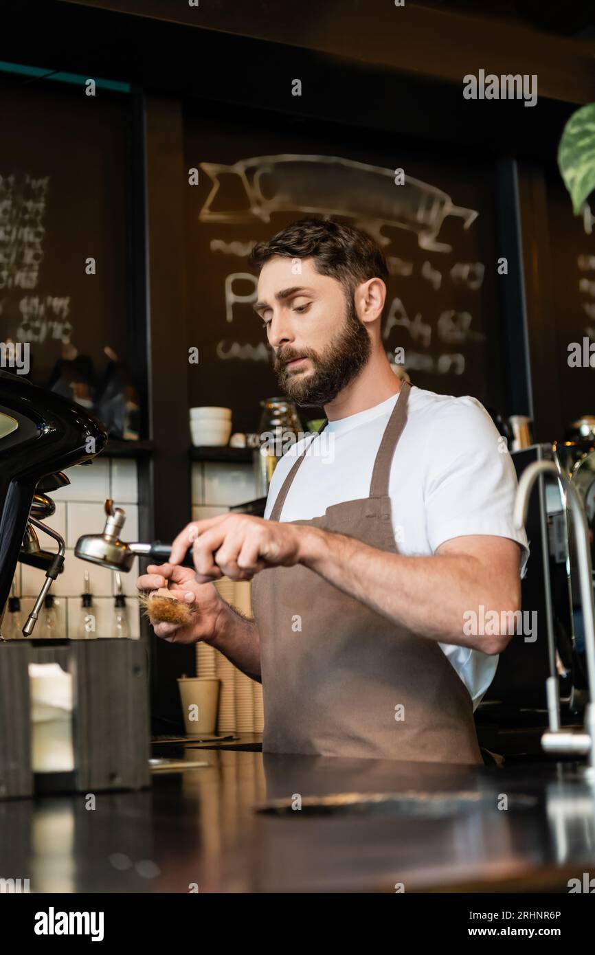 bearded barista in apron cleaning coffee machine holder with brush in