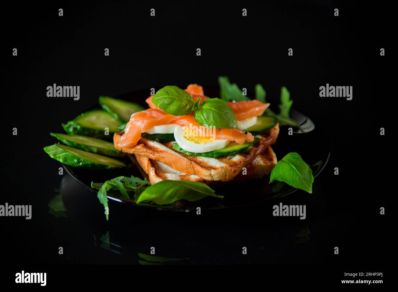 Cooked sandwich with lightly salted red fish, cucumbers and boiled eggs with herbs, isolated on a black background. Stock Photo