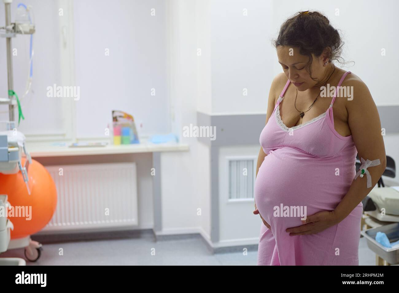 Middle aged multi-ethnic pregnant woman, birthing mother with painful contraction, at hospital ward, touching belly and looking down. Childbirth proce Stock Photo