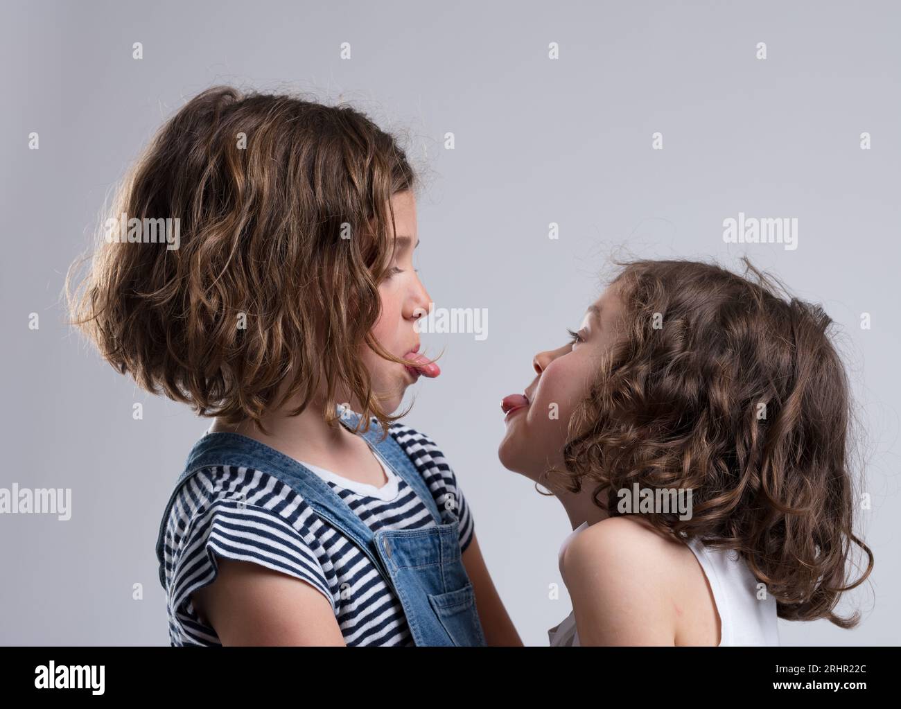 Two young girls, aged 5-7, teasingly stick out their tongues. Likely friends or sisters, they face each other making faces, dressed casually. They lau Stock Photo