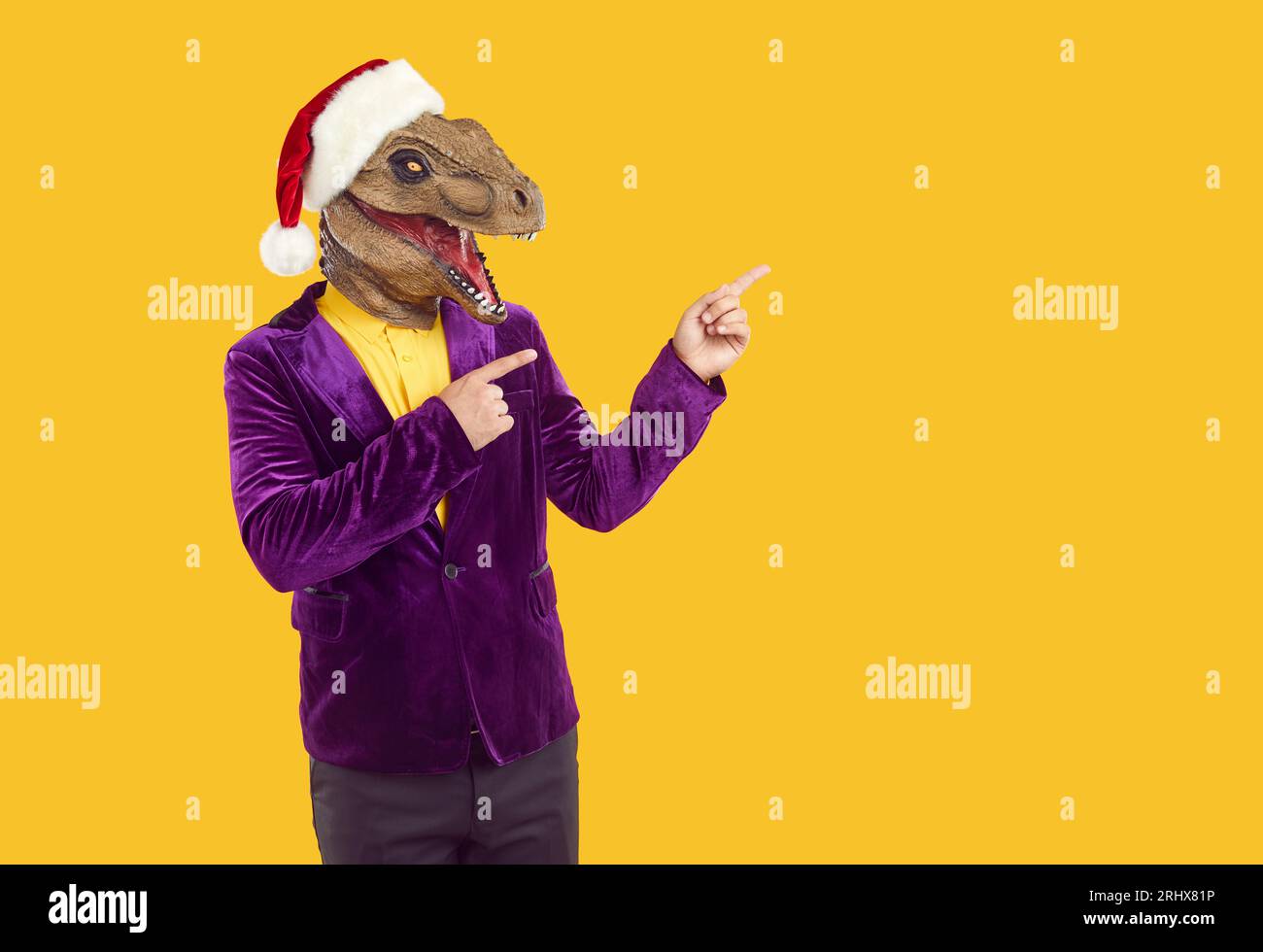 Funny man in dinosaur mask and Christmas hat points fingers to side on yellow background Stock Photo