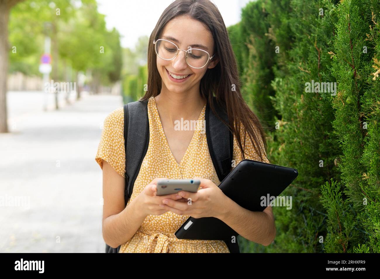 Smiling young female in young dress and eyeglasses browsing smartphone while standing with backpack and tablet against modern building and green bush Stock Photo