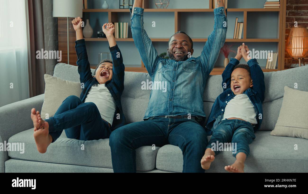 Excited African American happy family father and two siblings boys kids children fans watch TV sport game championship celebrate goal television team Stock Photo