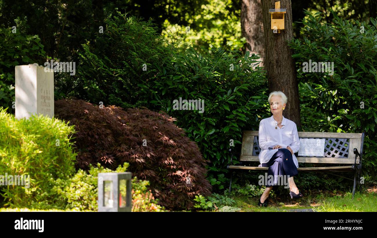 Cologne, Germany. 18th Aug, 2023. Dorothee Fiedler, initiator of a petition to preserve the private benches at Melaten Cemetery, sits on a bench at her son's grave at Melaten Cemetery. This summer, Cologne has a topic that is stirring up tempers: should 'wild benches' in the Melaten cemetery for celebrities be removed, or would that be heartless? (to dpa KORR 'The great Cologne cemetery bench debate' from 20.08.2023) Credit: Rolf Vennenbernd/dpa/Alamy Live News Stock Photo