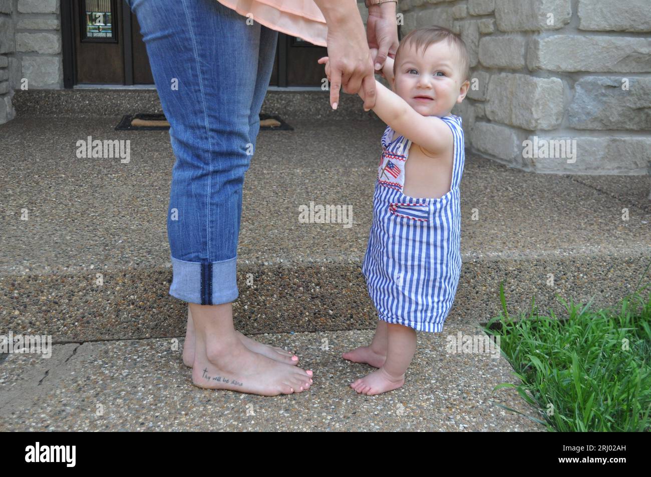 Mom holding baby's hand. Baby is standing with mom's help. Mom is barefoot. Happy baby standing. Scripture tattoo. Baby in American flag clothes. Stock Photo