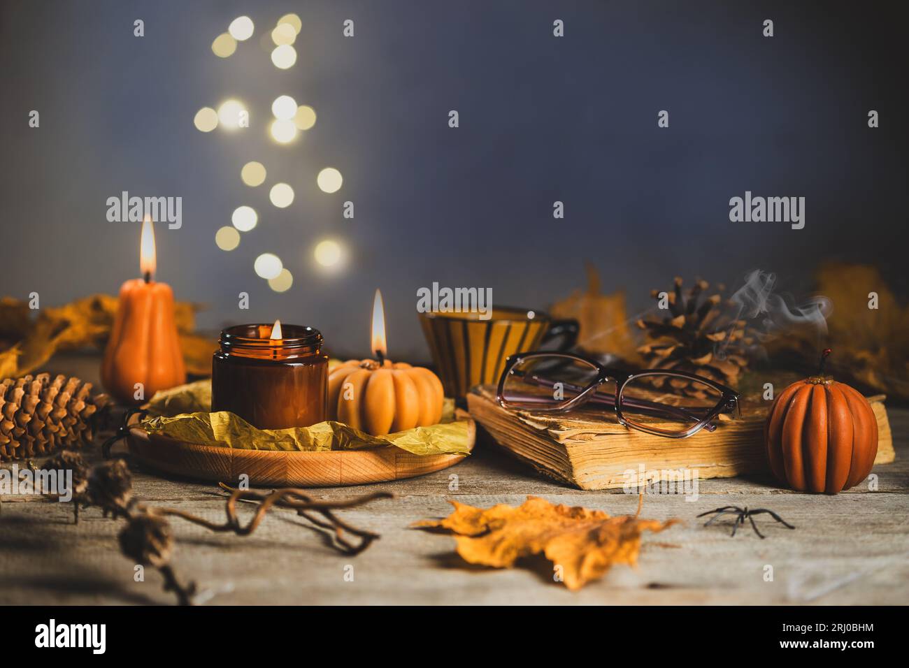 Halloween Still Life on the Wooden Table. Pumpkin Shaped Candles with dry Cones and Leaves, Old Book and Glasses. Bokeh Lights on the Background Stock Photo