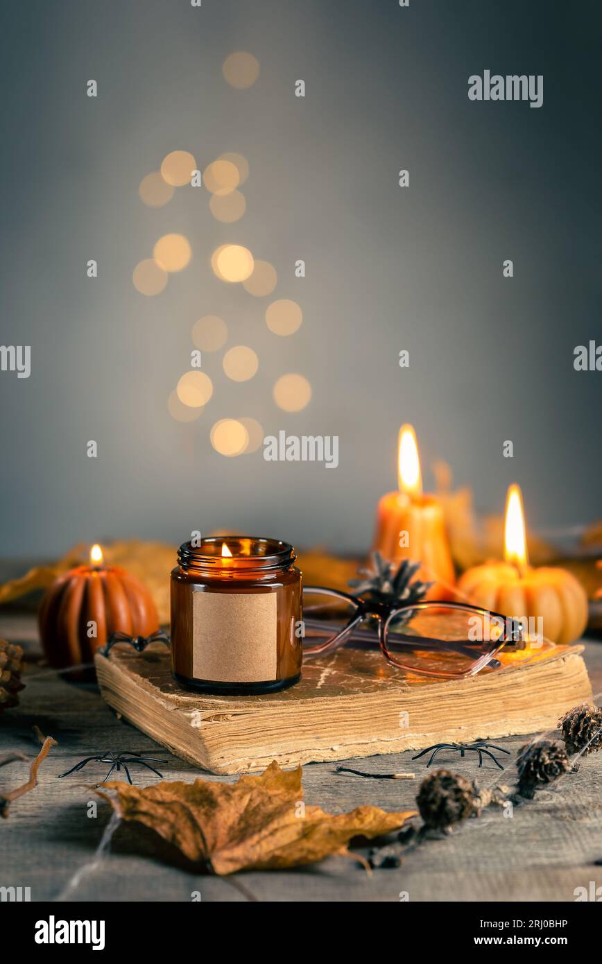 Halloween Still Life with Soy Wax Candle in the Dark Amber Glass with Empty Label for Mock Up. Pumpkin Shaped Candles with Cones, Leaves, Old Book and Stock Photo