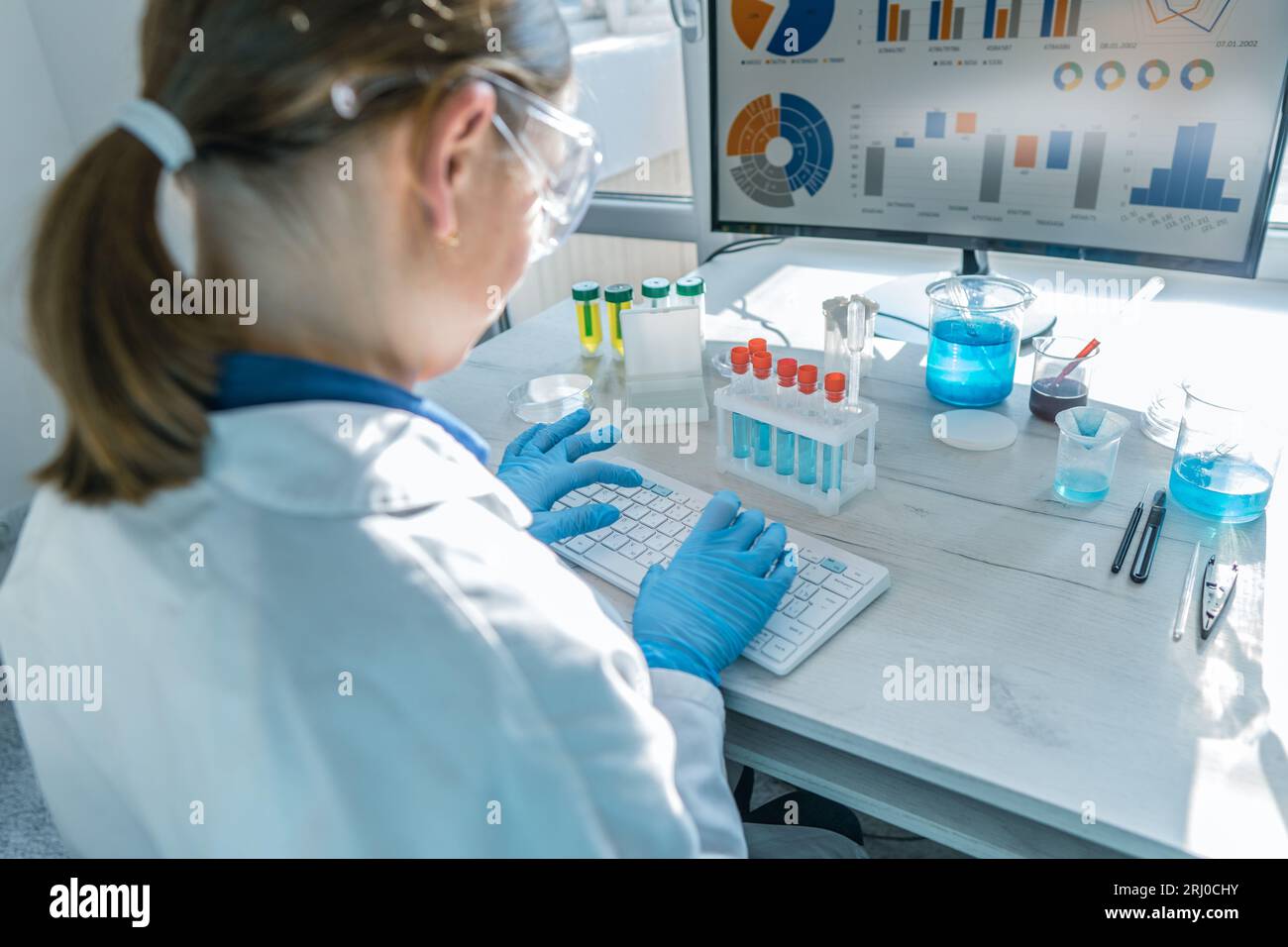 A female researcher sits at a workplace in a laboratory, behind a personal computer monitor. Against the background research statistics. Pharmaceutica Stock Photo