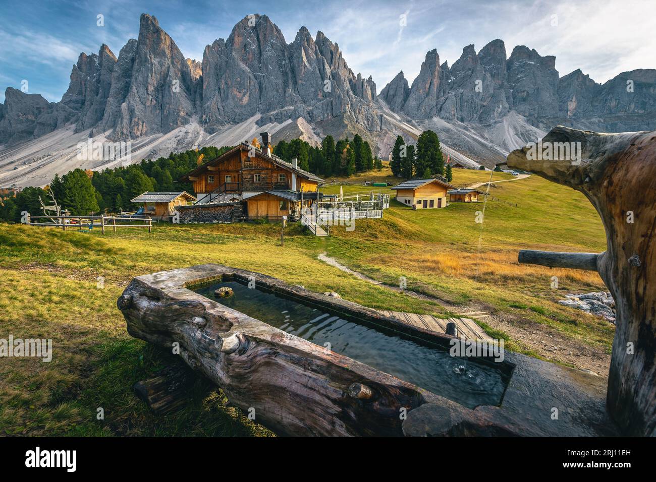 Small wooden well and cute chalets on the slope with spectacular high lacy peaks in background, Geisler - Odle mountain group, Alto Adige, Dolomites, Stock Photo