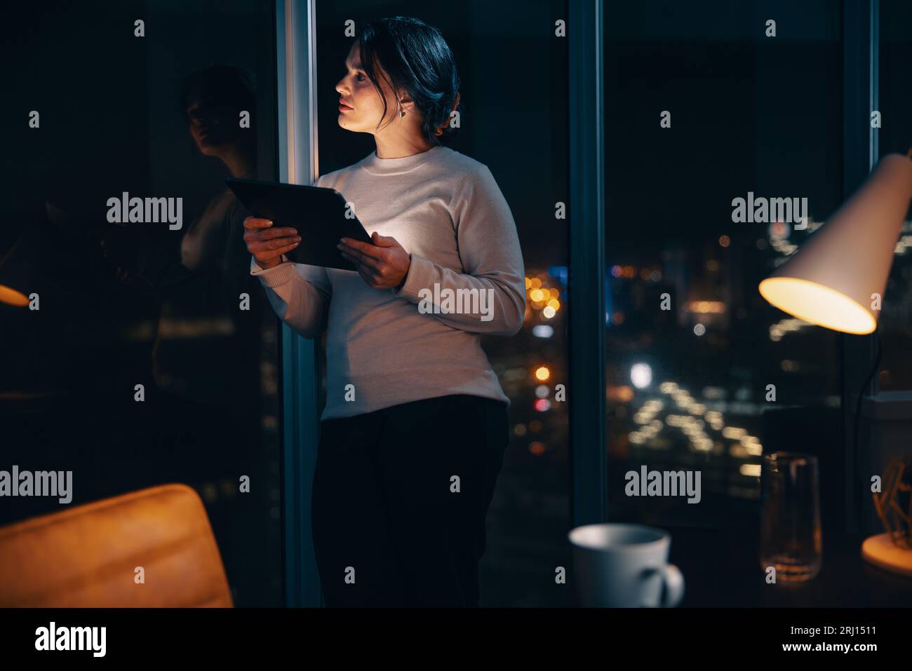 Thoughtful businesswoman using a digital tablet in her office at night, working overtime. She stands next to a window, focused and dedicated to her wo Stock Photo