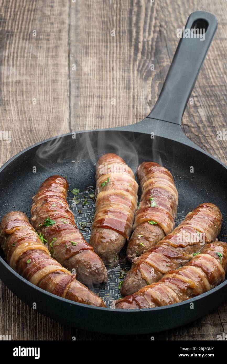 Appetizing  sausages wrapped in bacon and grilled in frying pan on wooden table Stock Photo