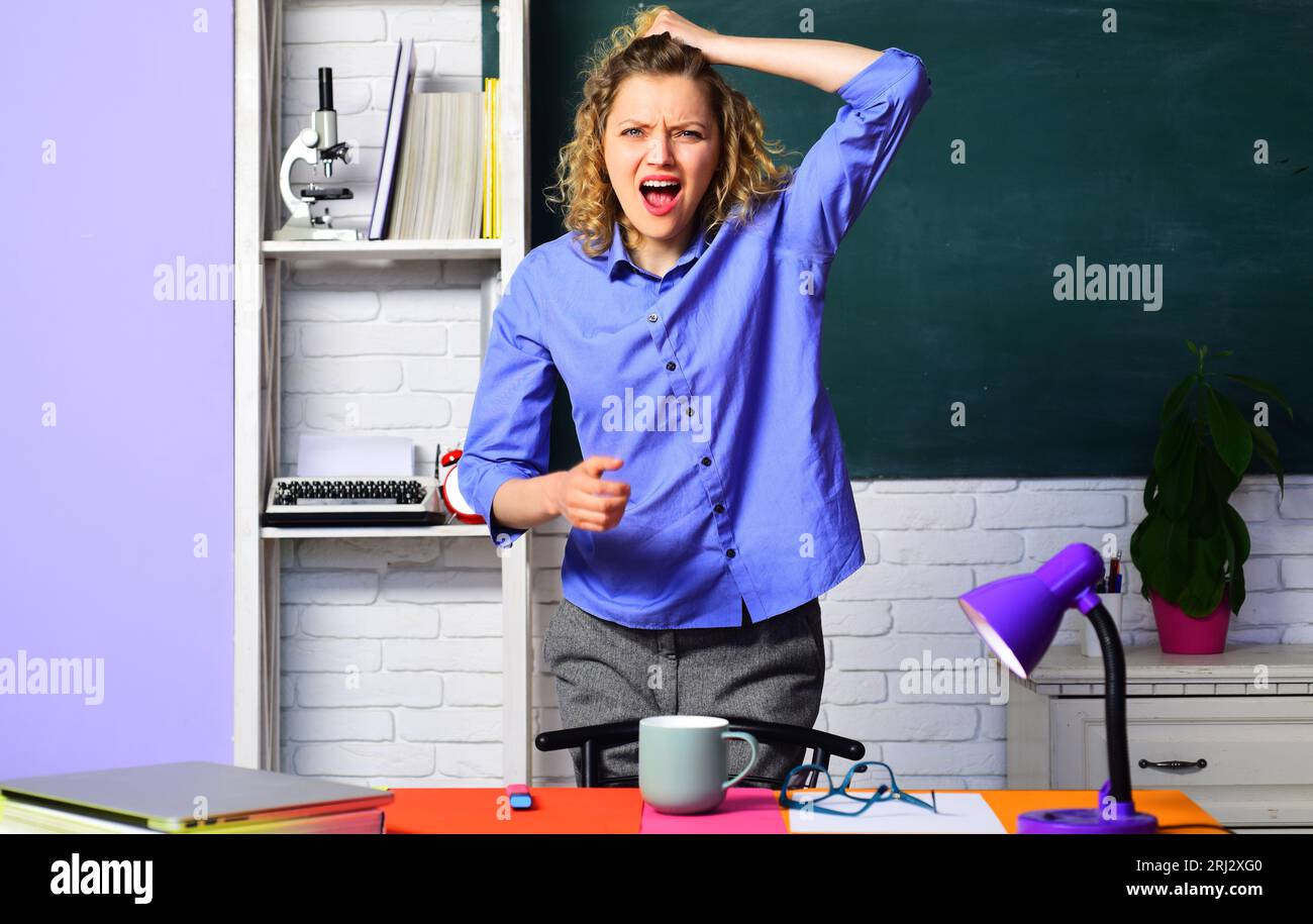 Screaming young teacher in classroom. Angry female teacher at university campus. Teachers job. Shouting student girl over near chalkboard. High school Stock Photo