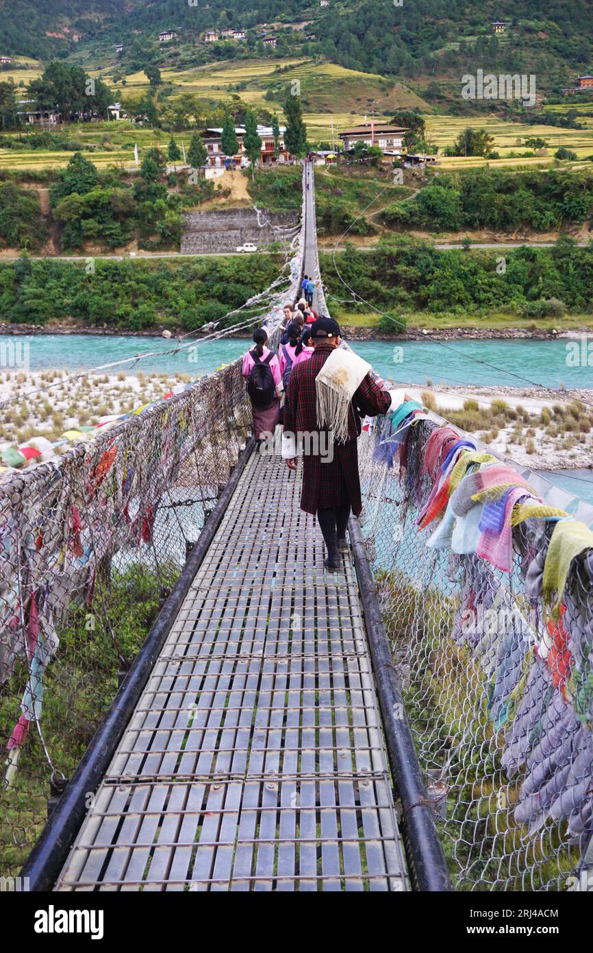 Bhutanese man, wearing traditional gho, walks behind a group of schoolgirls across the Punakha suspension bridge, spanning the Pho Chhu River, Bhutan Stock Photo