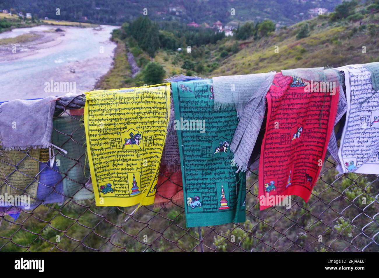 Colorful cloth prayer flags hang from a suspension bridge railing. The Pho Chhu River is visible in the valley beyond, near Punakha Dzong, Bhutan Stock Photo