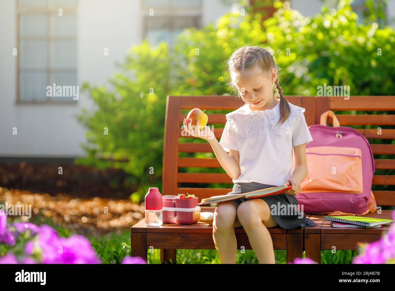 Pupil with lunch-box. Girl with backpacks is eating fruit near building ...