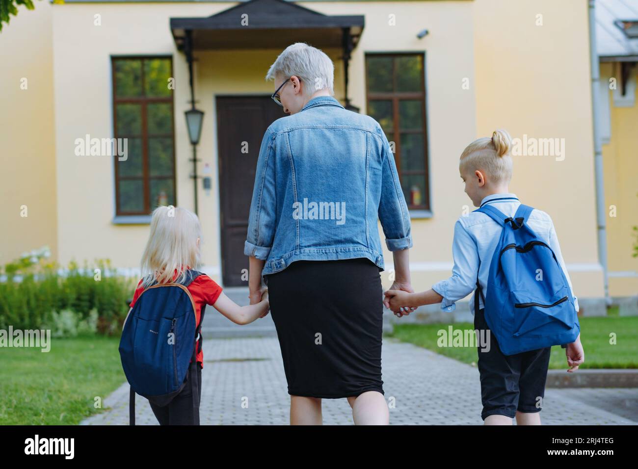 mother accompanies children to school holding hands. Caucasian blond boy with hairs tied up in ponytail and little girl going to school with mom. Imag Stock Photo