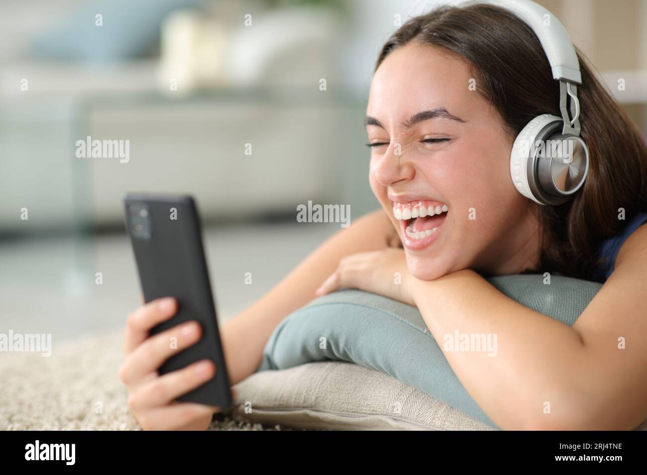 Happy woman wearing headphone watching videos laughing loud lying on the floor at home Stock Photo