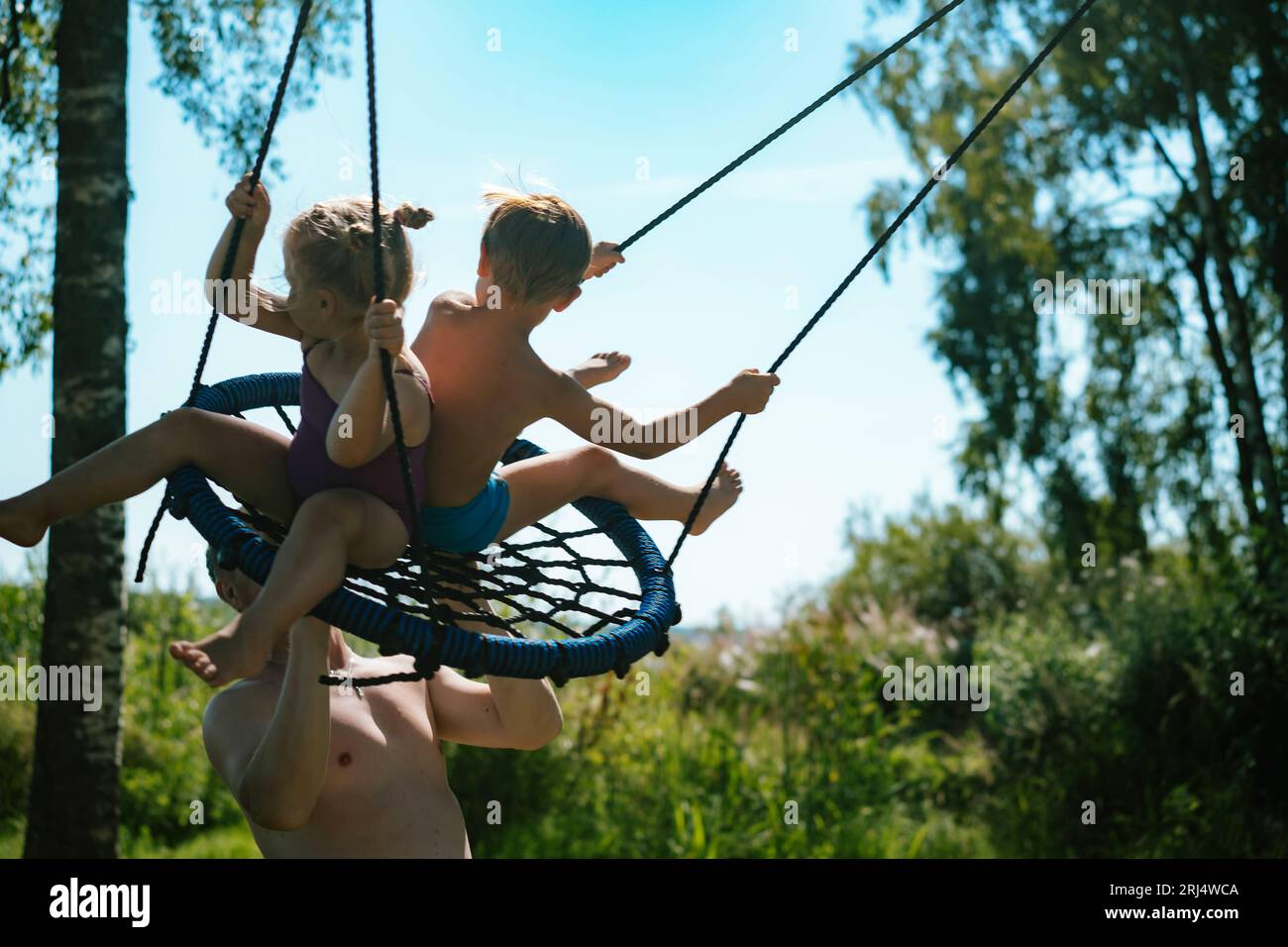 Father pushing children sitting on swing at playground. Summer in the nature. Quality time with family on vacation. Image with selective focus Stock Photo