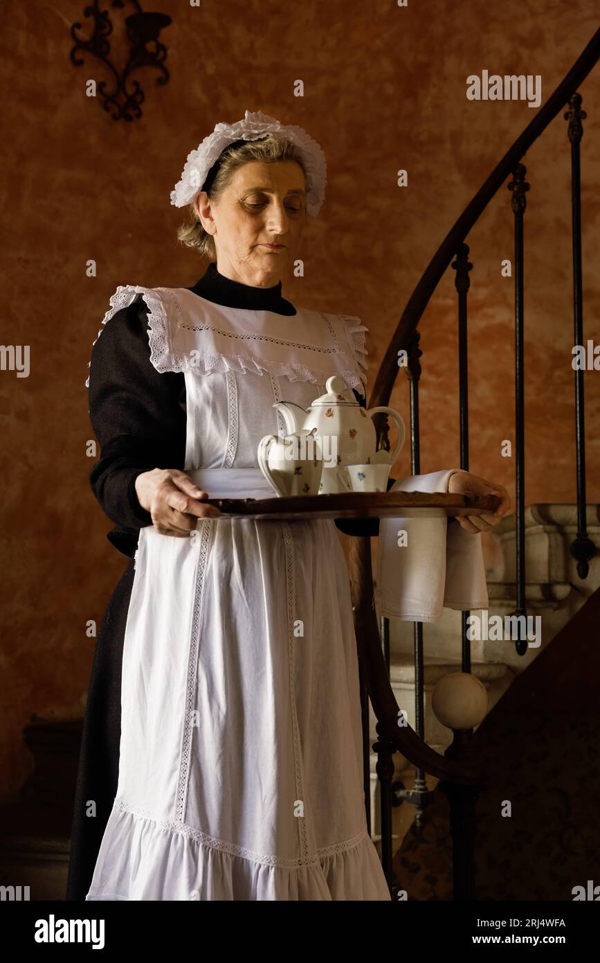 Victorian maid or servant in black dress, lace cap and white apron working in a 19th century interior Stock Photo