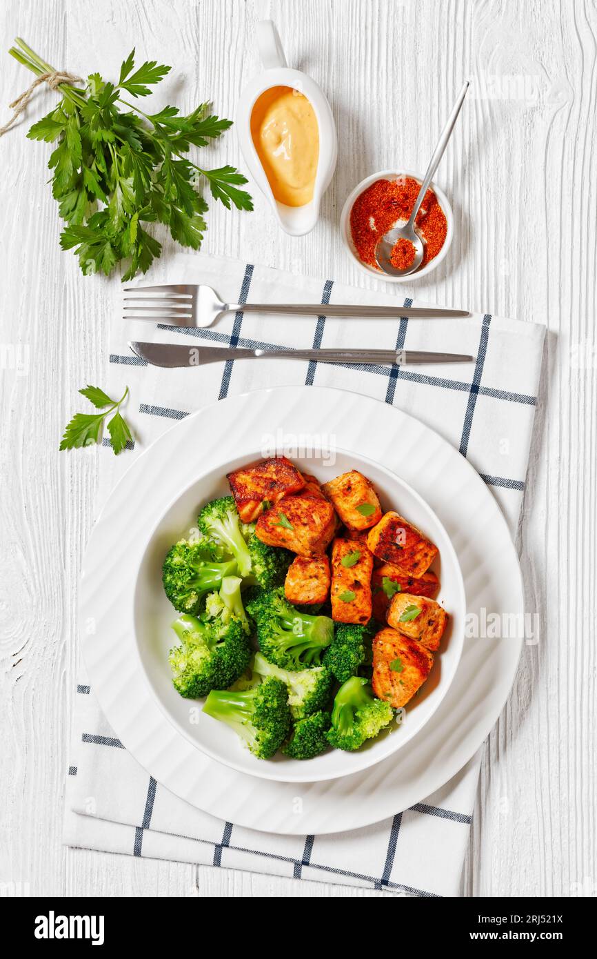 grilled salmon bites with steamed broccoli florets in white bowl on white wood table with sriracha mayo bang bang sauce, vertical view from above, fla Stock Photo