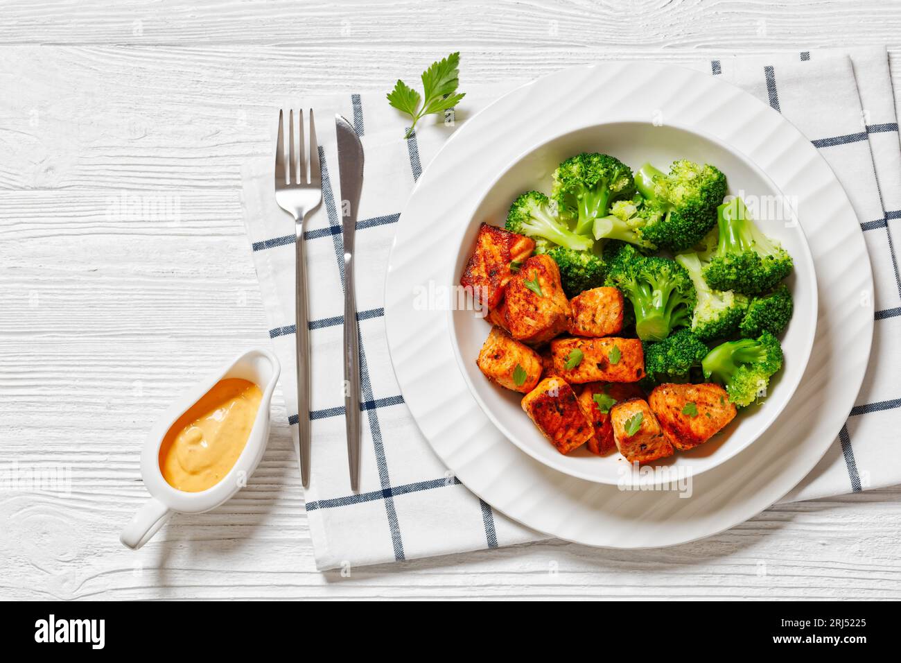 baked salmon bites with steamed broccoli florets in white bowl on white wood table with sriracha mayo bang bang sauce, horizontal view from above, fla Stock Photo