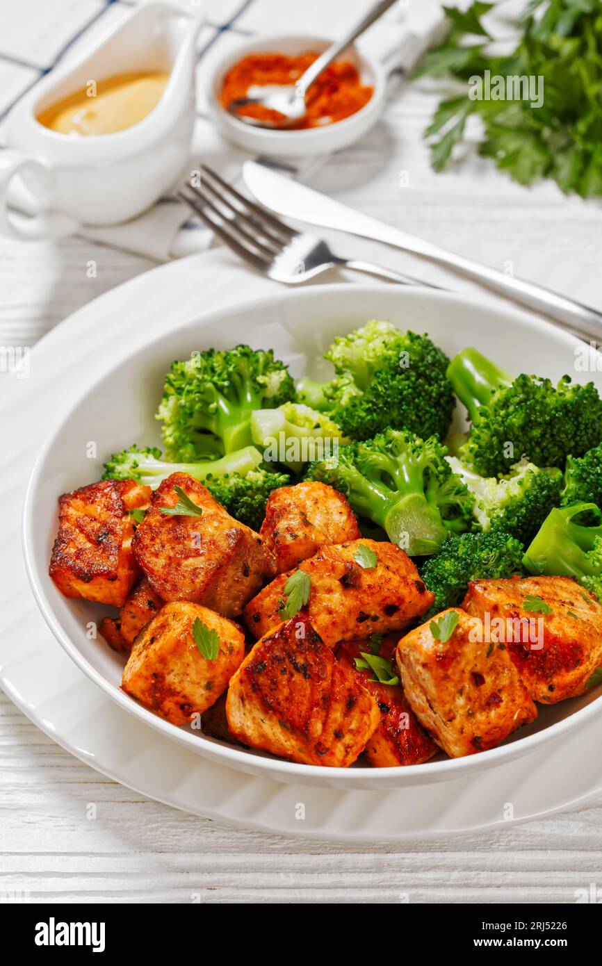 broiled salmon chunks with steamed broccoli florets in white bowl on white wood table with sriracha mayo sauce, vertical view from above, close-up Stock Photo