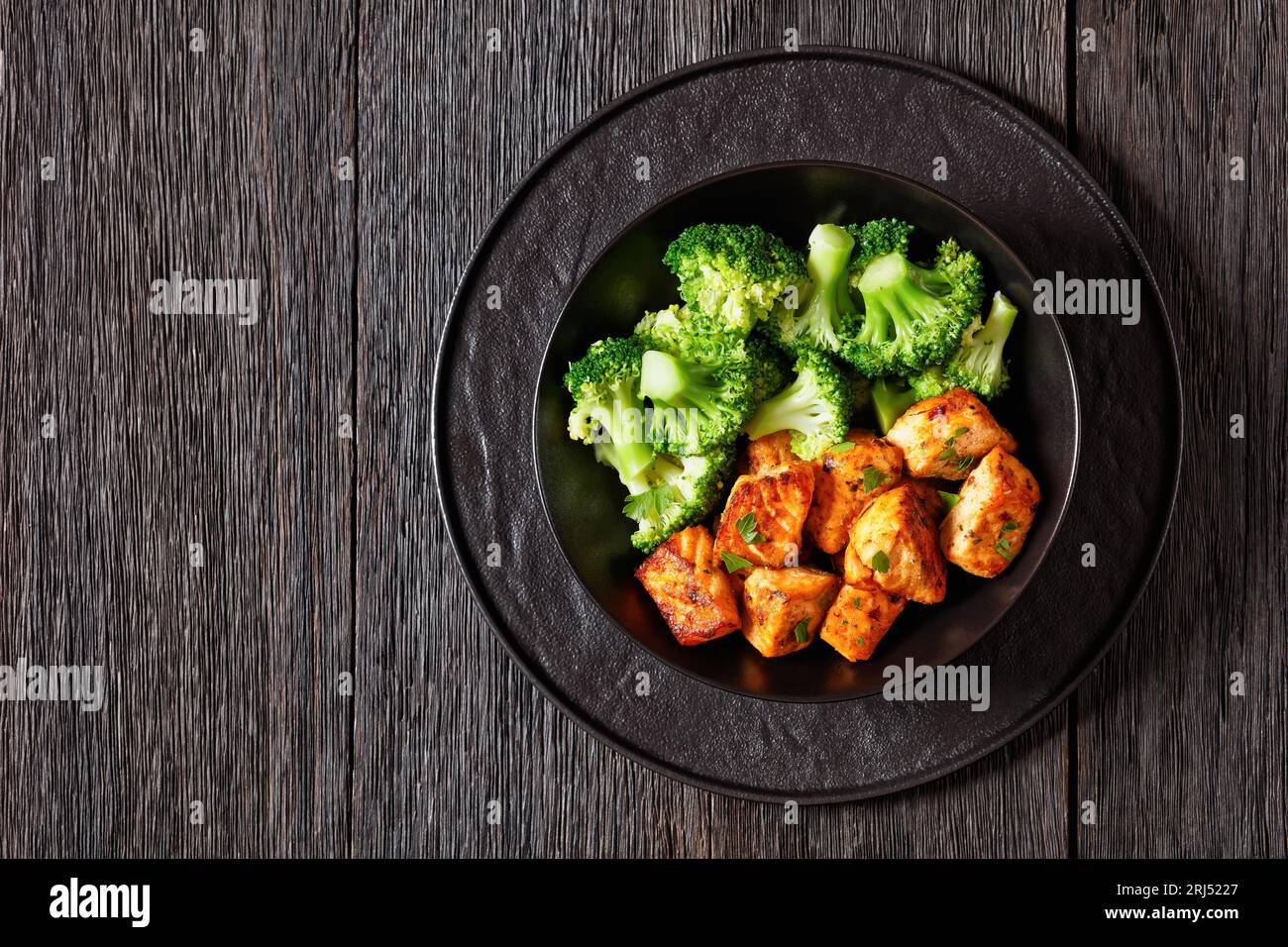 baked salmon chunks with cooked broccoli florets in black bowl on dark wooden table with sriracha mayo sauce, horizontal view from above, copy space Stock Photo