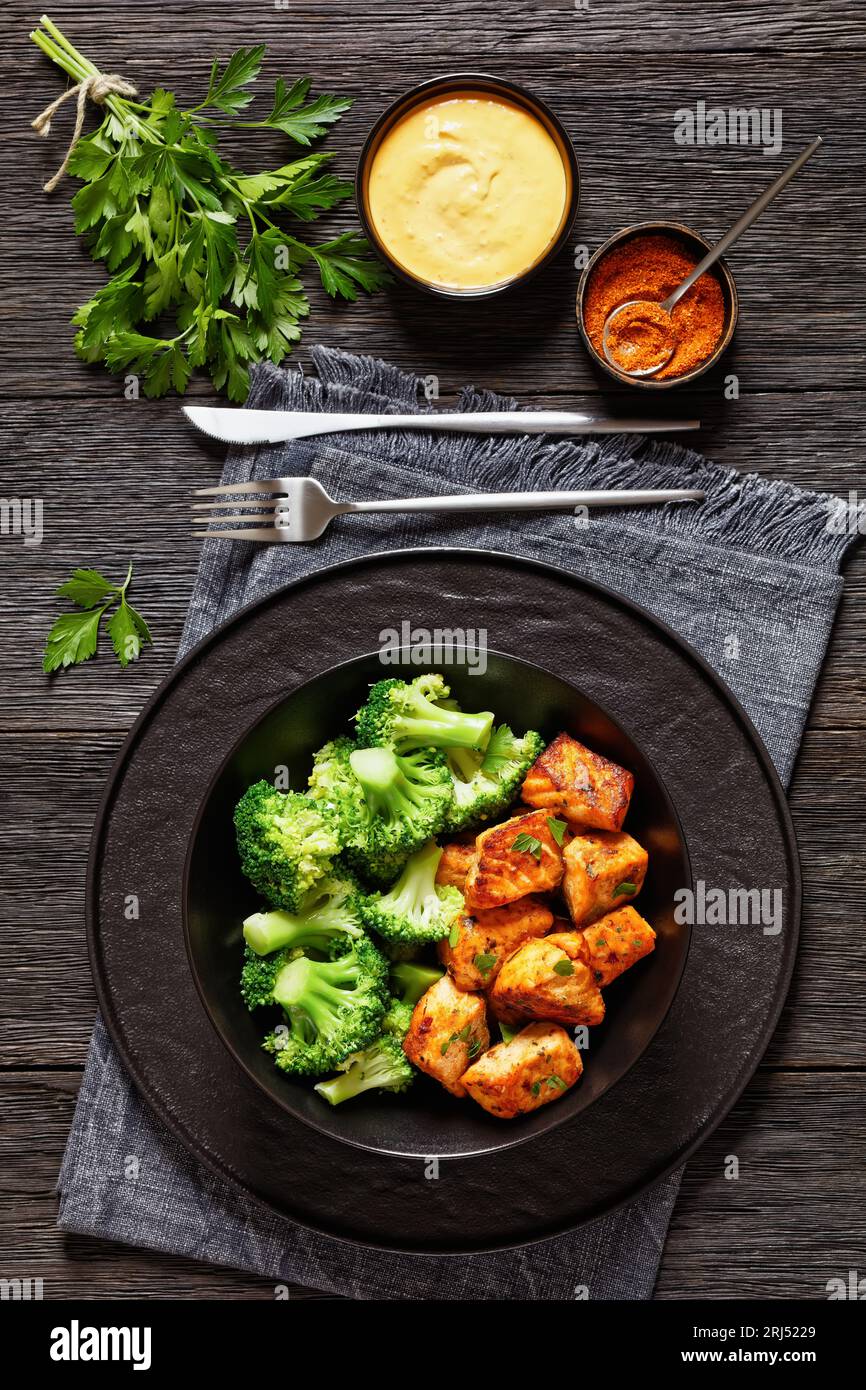 baked in oven salmon bites with steamed broccoli florets in black bowl on dark wooden table with sriracha mayo sauce, vertical view from above Stock Photo