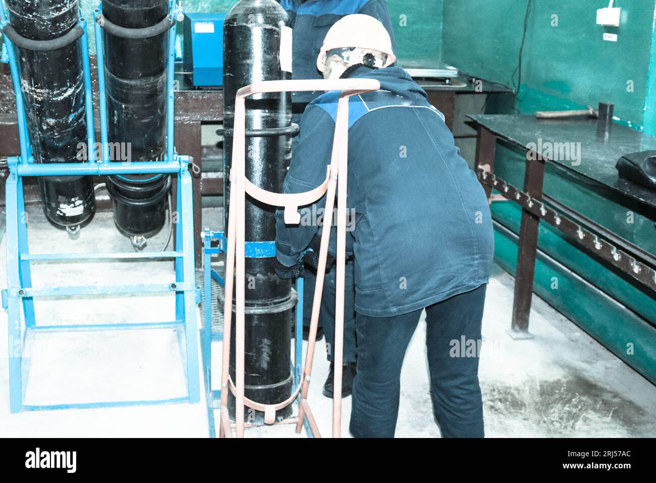 A worker in a helmet is loading a gas oxygen cylinder onto a trolley at an industrial plant. Stock Photo