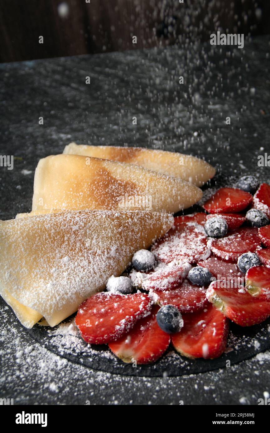 photo thin fried pancakes and lots of fresh berries sprinkled on top with powdered sugar on a dark background large Stock Photo