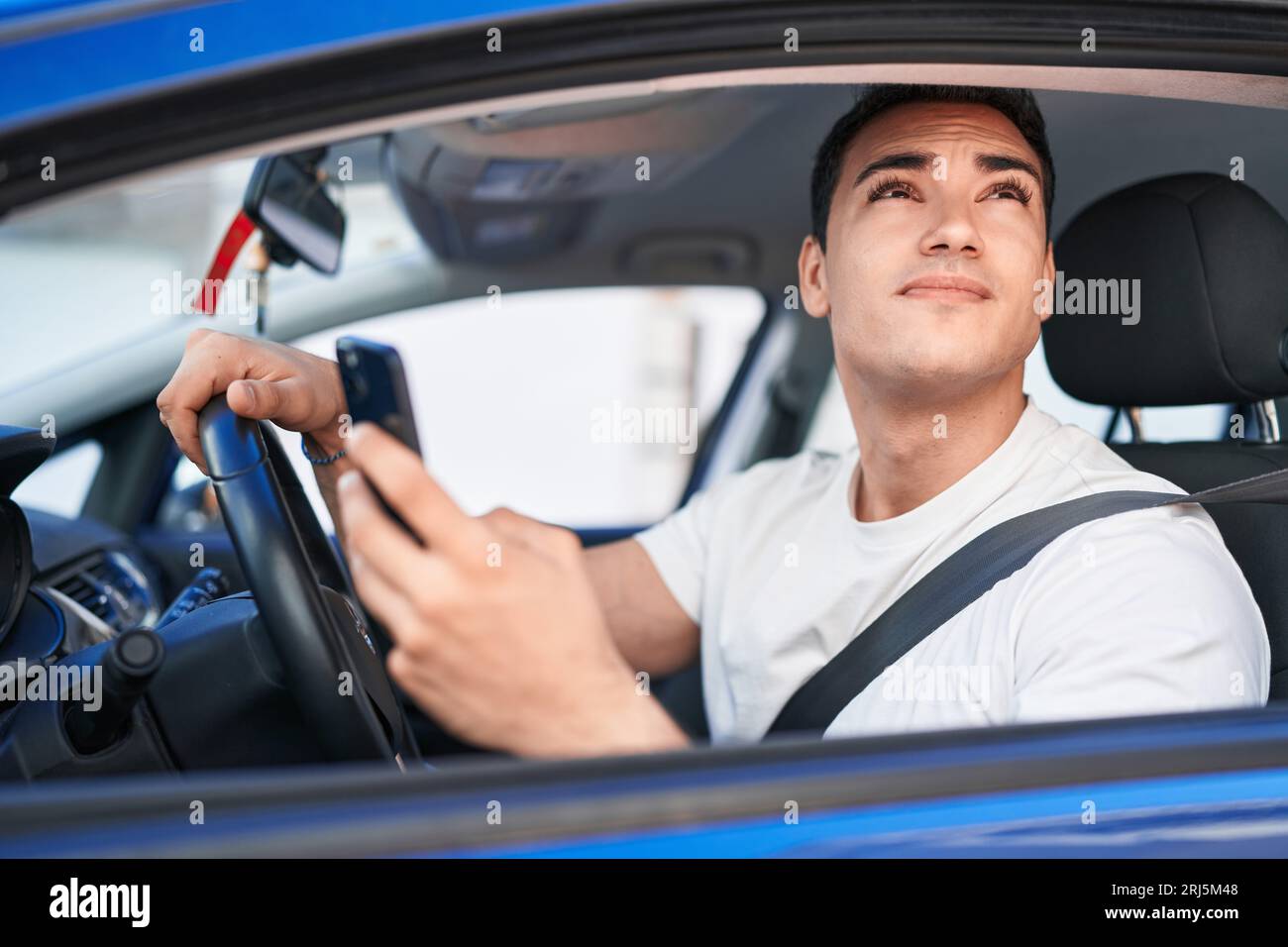 Young hispanic man using smartphone sitting on car at street Stock Photo