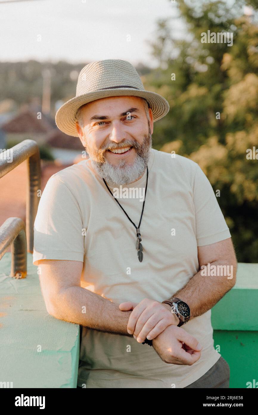 A mature man wearing a hat is sitting in a relaxed pose, facing the camera and smiling Stock Photo