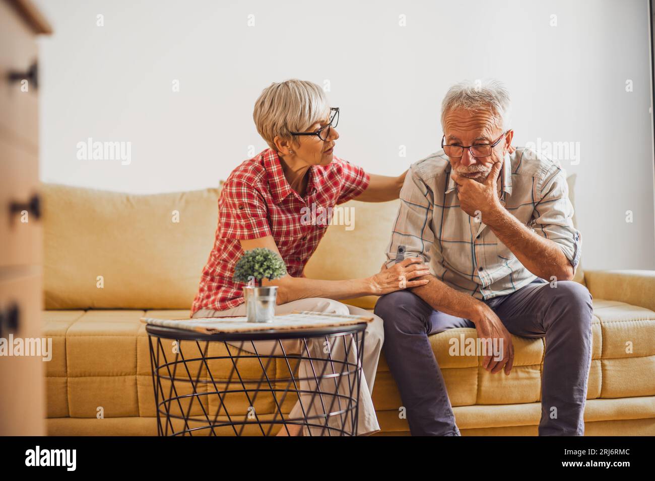 Senior couple sitting in living room. Man is sad and worried and woman is consoling him. Stock Photo