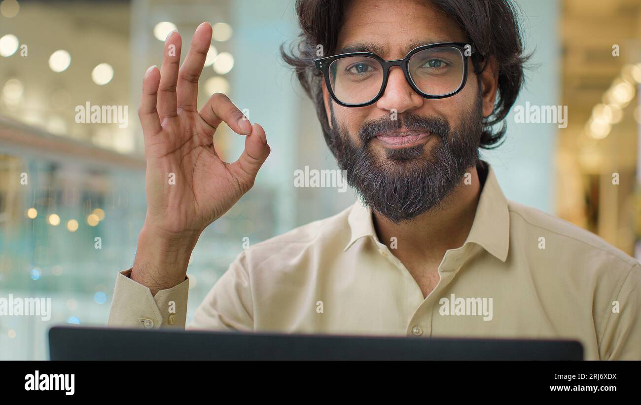 Indian Arabian bearded business man entrepreneur in glasses typing on laptop male professional businessman using computer sitting at office showing ok Stock Photo