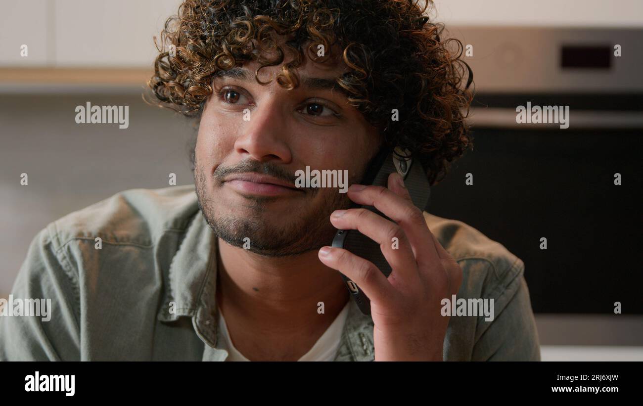 Cheerful Indian young man with curly hair talking phone in home kitchen. Happy friendly Hispanic guy talk with friends by smartphone Arabian male Stock Photo