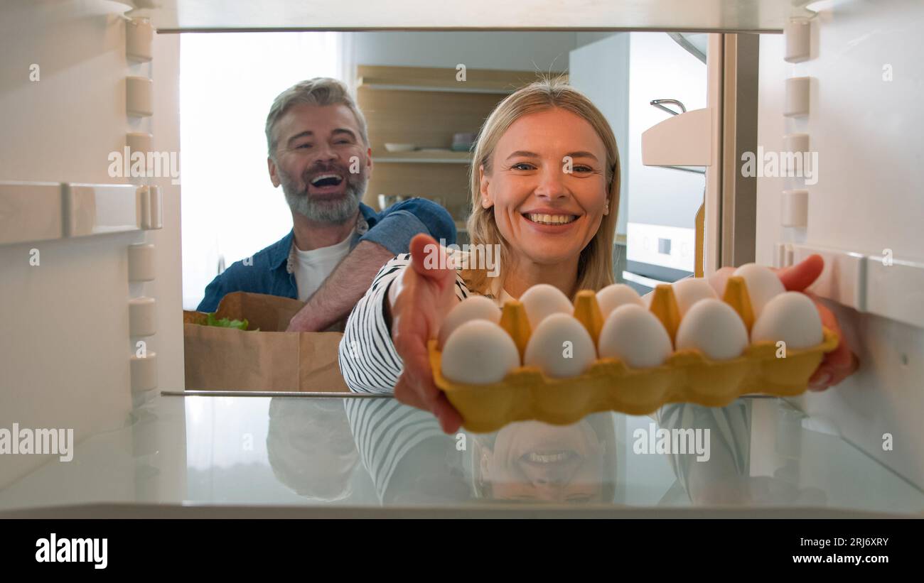 Point of view POV from inside refrigerator middle-aged couple spouses adult man and woman husband and wife taking vegetables food from paper bag Stock Photo