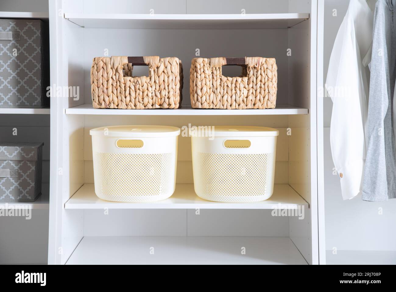 A white closet with multiple baskets on the shelves and several articles of clothing hanging from the closet rod Stock Photo