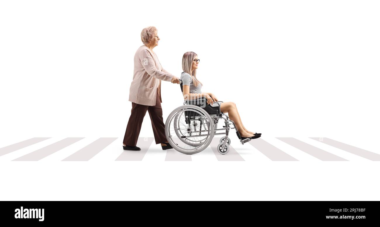 Full length profile shot of an older woman pushing a young woman in a wheelchair at a pedestrian crossing isolated on white background Stock Photo