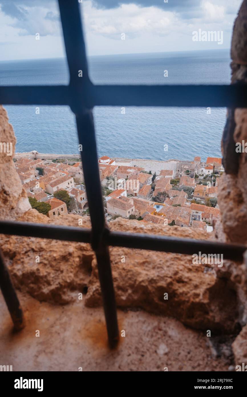 Ocean and City View through grid on rock stone wall in Monemvasia City, Cloudy Sky in Peninsula Peloponnese, Perfect Greece Scenery, Upright Format Stock Photo