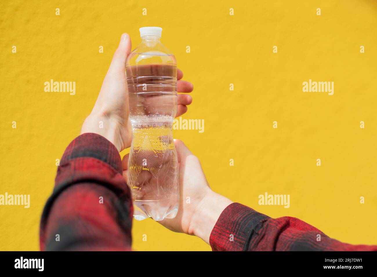 Close-up Unrecognizable man in a red shirt holds in his hands a Transparent Water Bottle in front of a yellow wall background Stock Photo