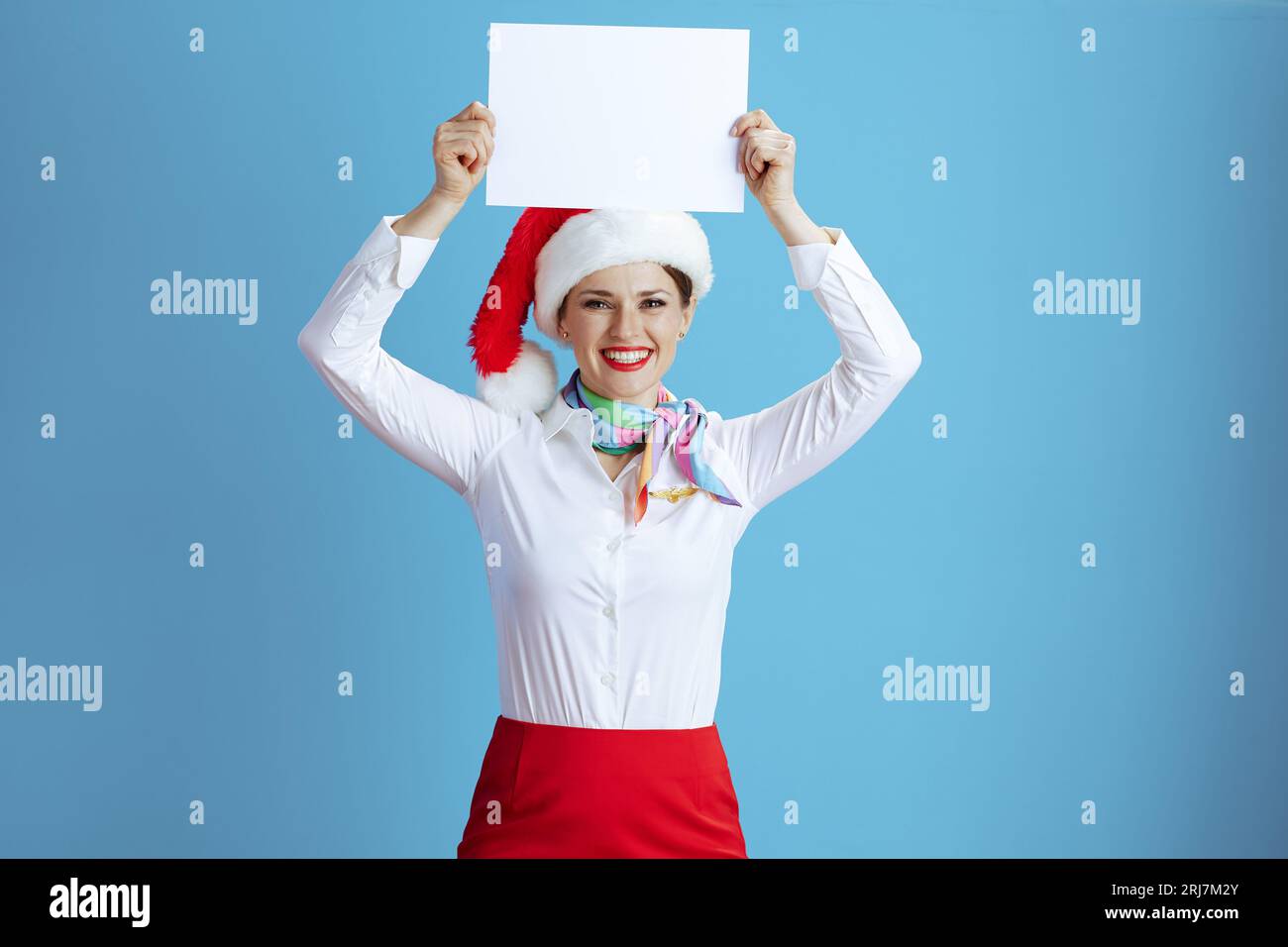 smiling modern female stewardess isolated on blue background in uniform with Santa hat showing blank a4 paper sheet. Stock Photo