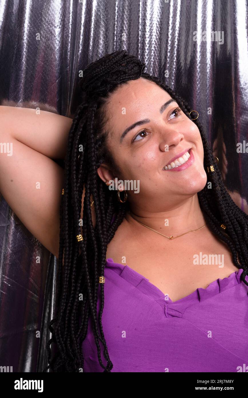 Beautiful woman with braids in her hair wearing lilac-colored outfit with her arms behind her head. Against silver colored background. Positive and ha Stock Photo