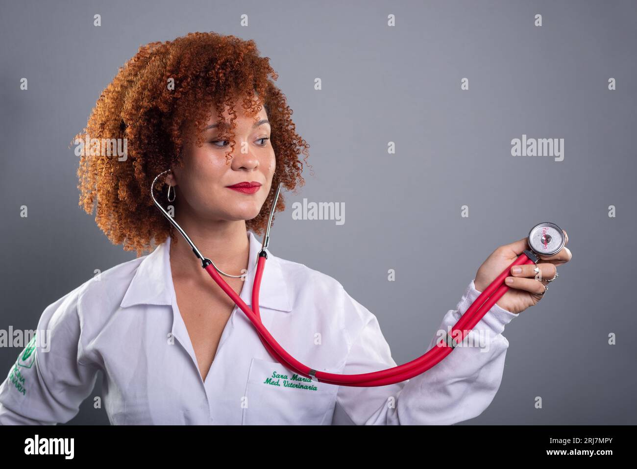 Veterinarian woman, pretty with red hair, showing a stethoscope to the camera in a white uniform. Isolated on gray background. Stock Photo