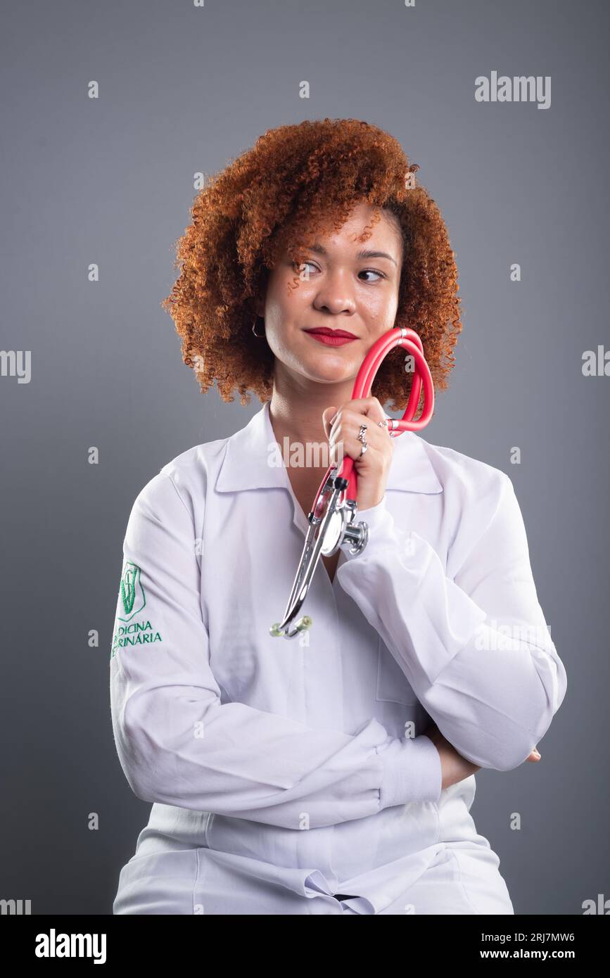 Portrait of a female veterinarian with red hair, wearing a white uniform and holding a stethoscope close to her face. Animal care. Isolated on gray ba Stock Photo