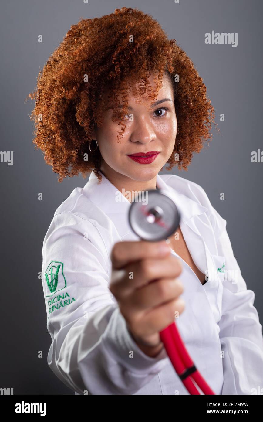 Portrait of a female veterinarian with red hair, wearing a white uniform and holding a stethoscope close to her face. Animal care. Isolated on gray ba Stock Photo
