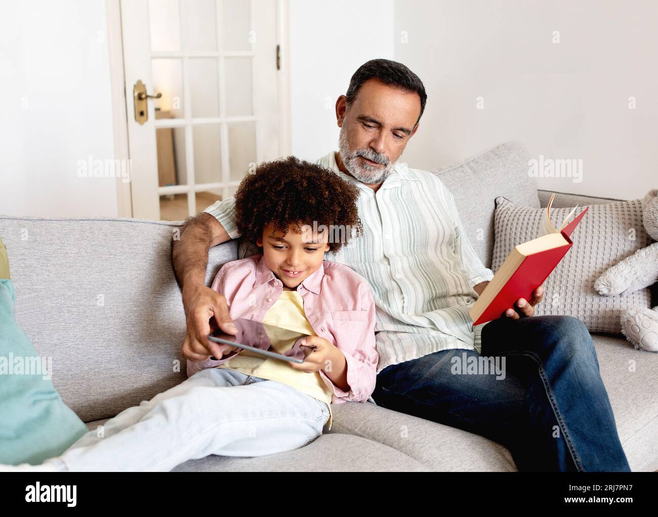 Hispanic Grandson Using Tablet Computer While Grandpa Reading Book Indoor Stock Photo