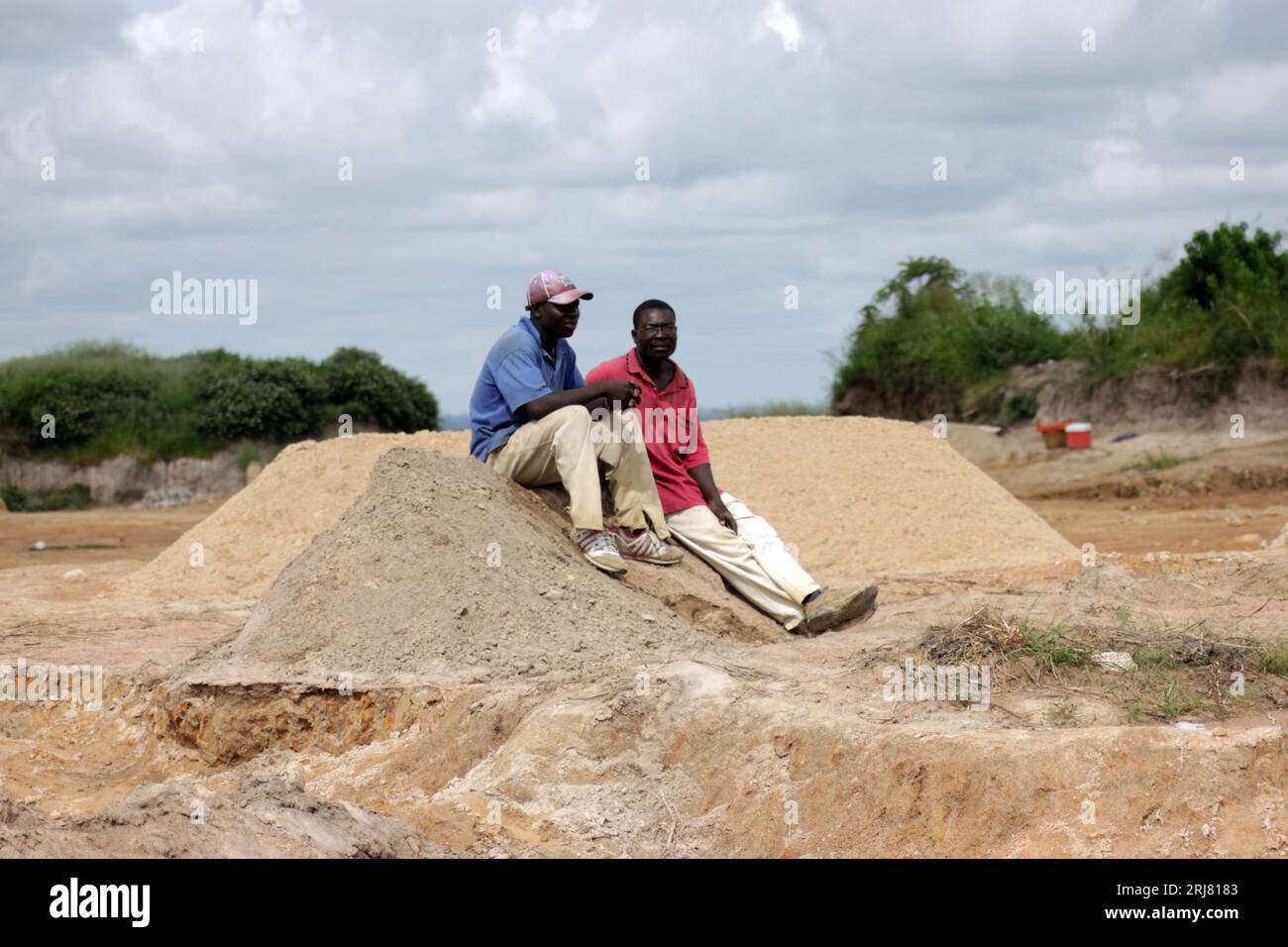 Two men are sitting on top of a mound of sand as they rest from digging for sand. Sand mining is illegal in most parts of the country. Harare, Zimbabwe. Stock Photo