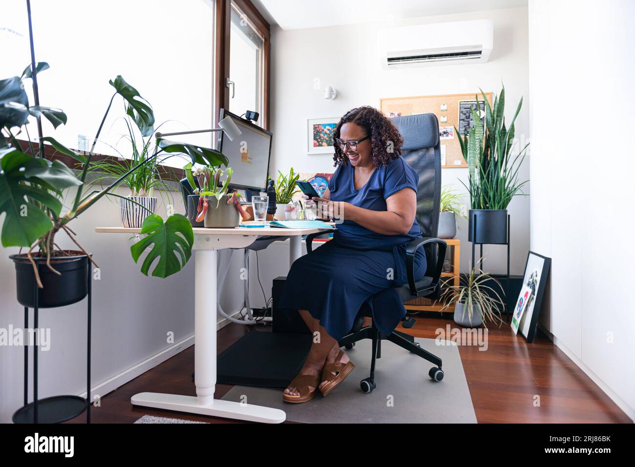Beautiful oversize black woman smiling while looking into her mobile screen in her home office decorated with plants Stock Photo