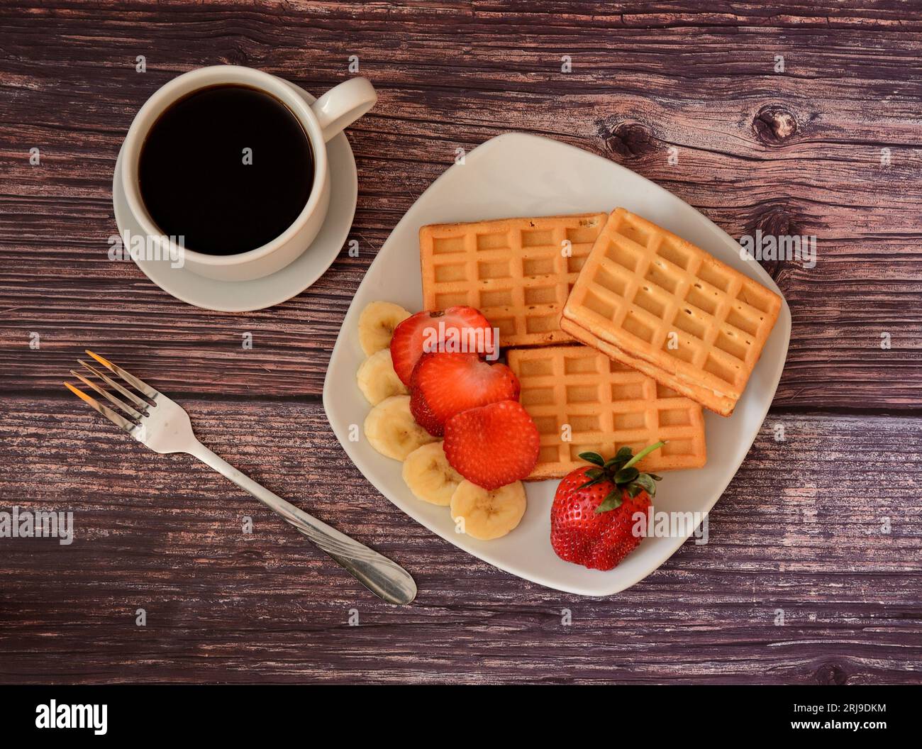A cup of black coffee on a saucer, a fork and a plate with fresh Viennese waffles with strawberries and ripe banana slices on a wooden table. Top view Stock Photo