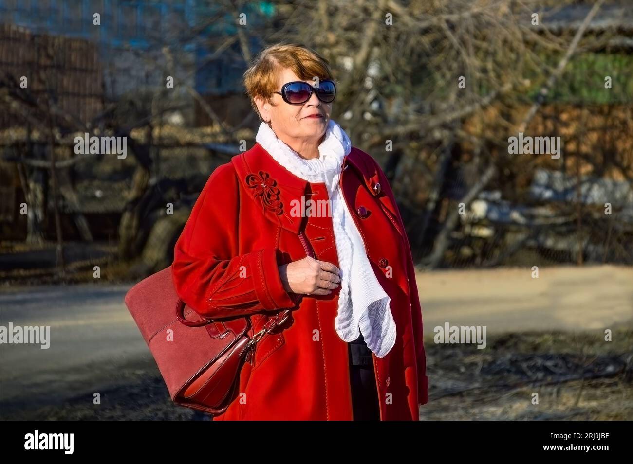 Portrait of stylish 67 year old woman in a bright red coat with a burgundy bag, sunglasses and white scarf on a spring day Stock Photo